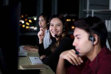 Mutlu gülümseyen Asyalı kadın çağrı merkezinde çalışıyor. Gece yarısı takım arkadaşı ile çalışıyor. Gece vardiyası müşteri hizmetleri personeli mikrofon kulaklığıyla müşteri desteği için bilgisayar kullanıyor.