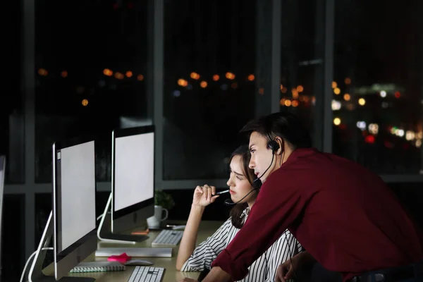 stock image Group of Asian night shift staffs with headphone look at desktop computer together for discussion, night shift worker team with hands-free phone work hard overtime at call center customer service care