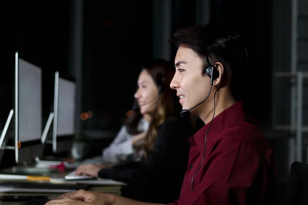 Happy smiling Asian man work at call center service consultant working with teammate colleague overnight, night shift customer service staff with microphone headset use computer for support customer