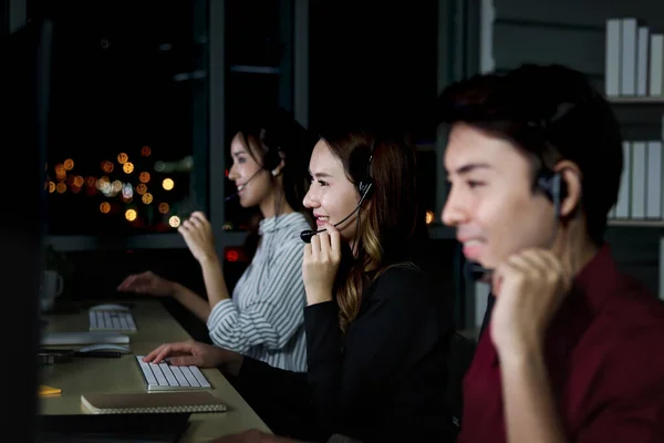 stock image Happy smiling Asian woman work at call center service consultant working with teammate colleague overnight, night shift customer service staff with microphone headset use computer for support customer