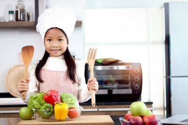 Asyalı gülümseyen kız portresi önlük takıyor ve aşçı şapkası mutfak tezgahında renkli taze sebzelerle duruyor salata malzemeleri, sağlıklı yemek pişiren tatlı küçük şef çocuk..