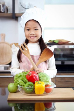 Asyalı gülümseyen kız portresi önlük takıyor ve aşçı şapkası mutfak tezgahında renkli taze sebzelerle duruyor salata malzemeleri, sağlıklı yemek pişiren tatlı küçük şef çocuk..