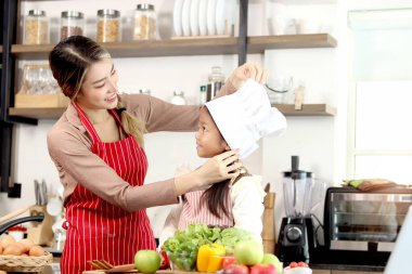 Mutfakta önlüğü ve şef şapkası olan mutlu Asyalı anne ve kız çocuğu için şapka takan anne, evde yemek pişiren aile şefi, anne ve çocuk vakit geçirip birlikte eğleniyorlar..