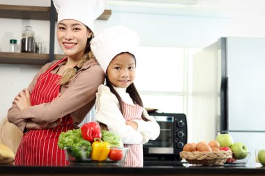 Mutlu gülümseyen Asyalı anne kızı portresi önlük şefi şapkası takıyor mutfakta kolları çapraz duruyor taze sebzelerle salata malzemeleri, tatlı şef ailesi sağlıklı yemekler pişiriyor..