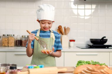 Mutlu gülümseyen Asyalı çocuk önlük ve şef şapkası takıyor, cam kasede yumurta karıştırıyor ve karıştırıyor, tatlı çocuk mutfakta yemek pişiriyor, sevimli aşçı çocuk tek başına yemek pişiriyor..