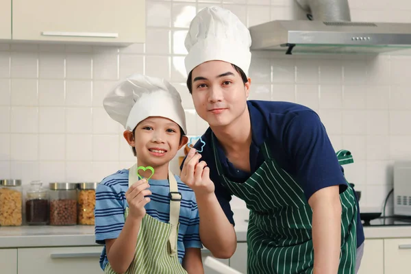 在厨房做饭时 戴着围裙和厨师帽的快乐的亚洲父亲和儿子拿着塑料饼干钳 可爱的家庭厨师做饭 父亲和男孩在一起消磨时间 — 图库照片
