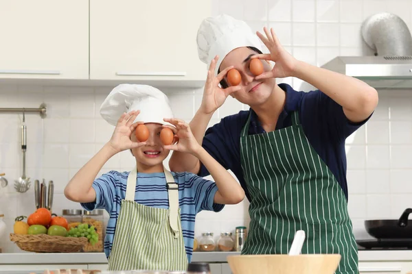 快乐的亚洲父亲和儿子 带着围裙和厨师帽 在厨房做健康的饭菜时拿着鸡蛋玩耍 可爱的家庭厨师做饭 爸爸和男孩在一起 — 图库照片