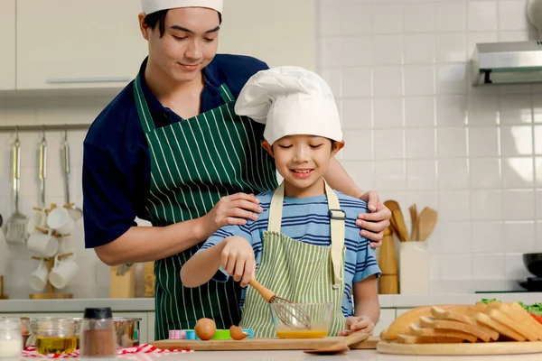 快乐地微笑着 带着围裙和厨师帽的亚洲男孩和男孩一起在厨房做饭 爸爸在帮助男孩搅拌鸡蛋 在玻璃碗里搅拌鸡蛋 可爱的家庭厨师做饭 — 图库照片