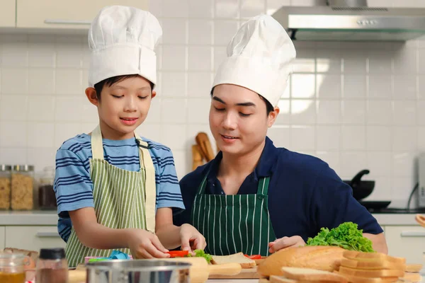 Mutlu gülümseyen Asyalı baba ve oğul önlüklü ve aşçı şapkalı çocuk mutfakta birlikte yemek pişirirken baba ve baba çocuk basit bir yemek hazırlarken çocuğa yardım ederken sevimli aile şefi yemek pişirirken..