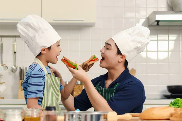 快乐的亚洲父亲和儿子孩子们 带着围裙和厨师帽一起在厨房做饭 爸爸和孩子们正在准备吃三明治 可爱的家庭厨师在做饭 吃得很开心 — 图库照片