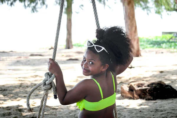 明るい色の水着を着て黒い巻き毛を持つ幸せな笑顔アフリカの女の子 ビーチの屋外でスイングに座って 晴れた日に外で遊んでいる美しい子供 子供のための夏の活動 — ストック写真