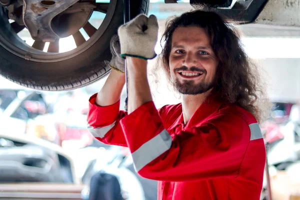 Kırmızı Üniformalı Uzun Saçlı Yakışıklı Tamirci Kaldırılmış Araçla Çalışıyor Garajda — Stok fotoğraf