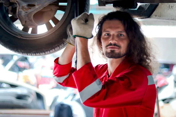 Kırmızı Üniformalı Uzun Saçlı Yakışıklı Tamirci Kaldırılmış Araçla Çalışıyor Garajda — Stok fotoğraf
