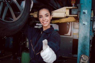 Güzel kadın oto tamircisi garajda kontrol tekerlerini kontrol ederken başparmağını bırakıyor, araba servisi teknisyeni kadın araba servisinde müşteri arabasını tamir ediyor, araç motorunu kontrol ediyor..