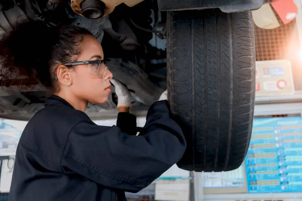 Beautiful female auto mechanic checking wheel tires in garage, car service technician woman repairing customer car at automobile service, inspecting vehicle underbody and suspension engine system.