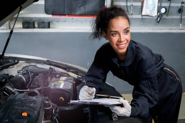 Beautiful female auto mechanic working in garage, car service technician woman holding file paper and checking car engine, repairing customer car at automobile service, inspecting vehicle engine.