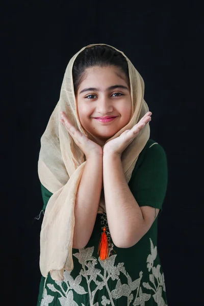 ヒジャーブと伝統衣装を着た美しい目を持つ愛らしい幸せな笑顔パキスタンのイスラム教徒の女の子 かなりかわいいですインドの子供立っていると暗い黒の背景に手で顎を休める — ストック写真