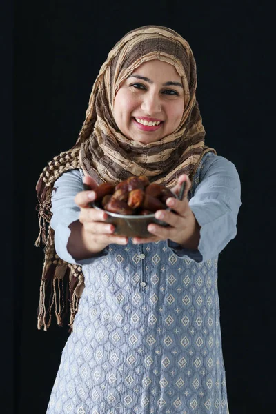 Portrait beautiful middle aged smiling Asian Muslim woman were hijab and traditional costume, hold date fruits, sweet and energy boost food mostly eating in Ramadan of Islam on dark black background.