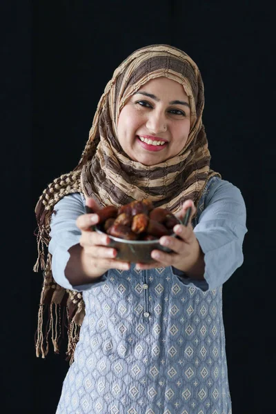 Portrait beautiful middle aged smiling Asian Muslim woman were hijab and traditional costume, hold date fruits, sweet and energy boost food mostly eating in Ramadan of Islam on dark black background.