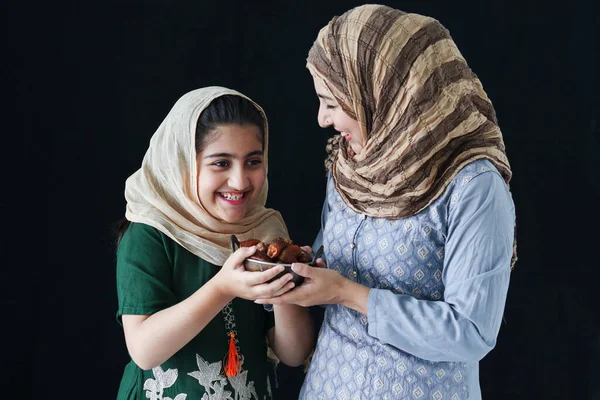 Beautiful Asian Muslim woman with daughter girl wear hijab scarf and traditional costume, hold date fruits together, sweet and energy boost food mostly eat in Ramadan of Islam on dark black background