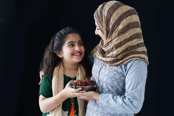 Beautiful Asian Muslim woman with daughter girl wear hijab scarf and traditional costume, hug each other, hold date fruits together, sweet food eating in Ramadan of Islam on dark black background.
