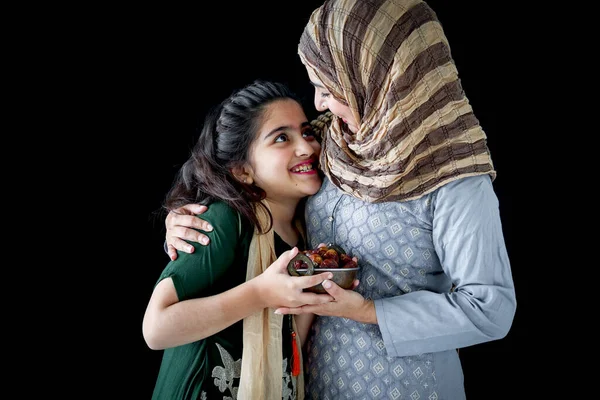 娘の女の子と美しいアジアのイスラム教徒の女性は ヒジャーブのスカーフと伝統的な衣装を着て 抱擁し 一緒に日付フルーツを保持し 暗い黒の背景にイスラムのラマダーンで甘い食べ物を食べる — ストック写真
