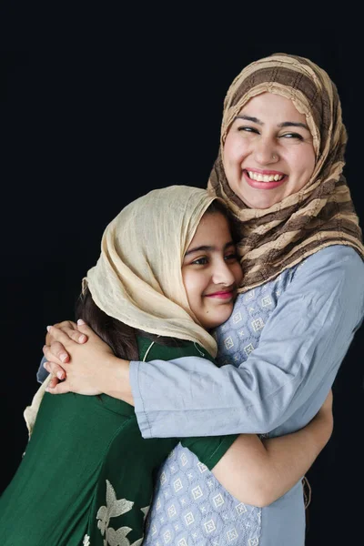 stock image Happy smiling Muslim girl with beautiful eyes wear hijab scarf and traditional costume hugging with her mother, warm love in family of mom and daughter, lovely Islamic family on dark black background.