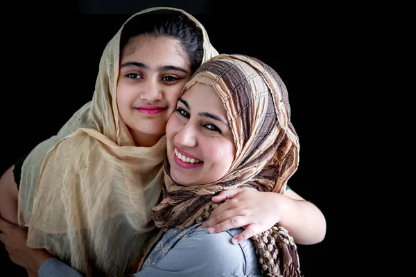 stock image Happy smiling Muslim girl with beautiful eyes wearing hijab scarf and traditional costume hugging with her beautiful mother, warm love in family of mom and daughter, lovely Islamic family on dark black background.