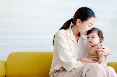 Mutlu gülümseyen bebek, sarı kanepede oturan anne kucağında oturuyor, anne küçük kızını öpüyor, ebeveyn tatlı bebeğe sarılıyor, aileyi seviyor, çocuğa bakıyor ve bakıyor..