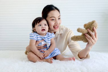 Anne oyuncak ayıyı tutuyor ve üzgün kızıyla oynamaya çalışıyor, üzgün bebek bebek oturma odasında oturuyor, anne kucağında ve küçük kızı kucaklıyor, ebeveyn evde bebeğe bakıyor, aile içinde sevgi var..