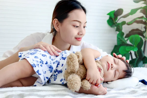 Cute toddle baby girl infant sleeping during hug teddy bear toy on bed, young beautiful mother looks at sleepy daughter kid, mom comforting sleeping kid, parent take care child at home, love family.