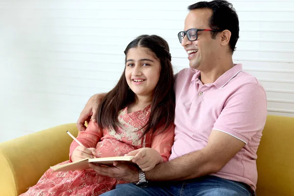 Cute Indian school student girl wears traditional dress sitting with father in living room and doing homework, dad teaching daughter kid at home, parent involvement in childhood education in family.