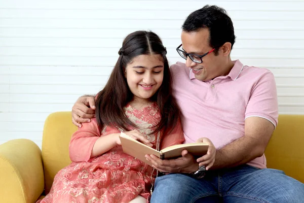 Cute Indian school student girl wears traditional dress sitting with father in living room and doing homework, dad teaching daughter kid at home, parent involvement in childhood education in family.