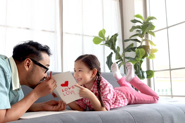 父亲节快乐 可爱的小女孩和年轻的爸爸躺在客厅的沙发上 制作并赠送手工制作的贺卡进行校准 可爱的家庭女儿和父亲在一起 — 图库照片