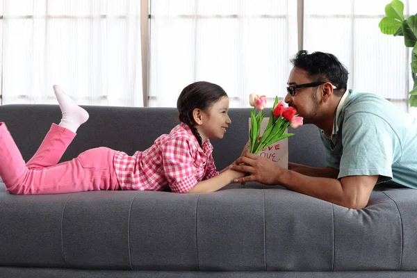 父亲节快乐 可爱的小女孩和年轻的爸爸躺在客厅的沙发上 把郁金香花送给她的爸爸进行校准 可爱的家庭女儿和父亲在一起 — 图库照片