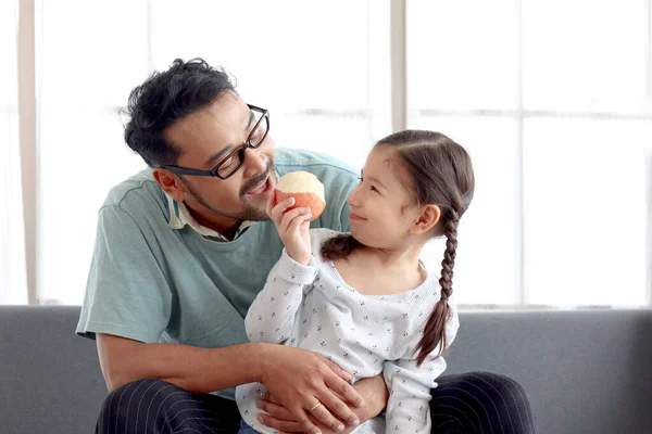 可爱的小女孩和她的爸爸坐在客厅的沙发上 一起玩 一起吃苹果 可爱的家人 爸爸和女儿一起呆在家里 照顾孩子 父亲节快乐 — 图库照片