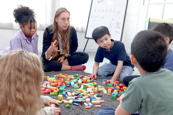 Group of primary school children study and play together in classroom with teacher, diverse students sit on floor while do activities and have fun, kid education, variety and diversity classroom.