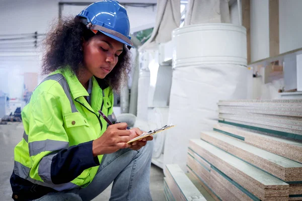 严重的非洲女工 头戴安全帽 穿着安全背心 在文件文件夹上书写 在家具厂检查木材原材料 数控木工 — 图库照片