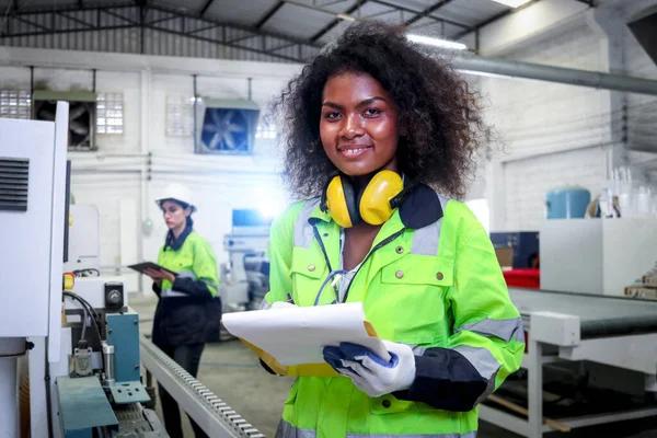 stock image African industrial woman worker with curly hair wears safety vest, writes on file folder document, female engineer check and maintenance machinery at CNC woodworking manufacturing furniture factory.