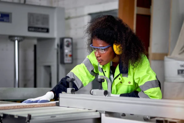 stock image African industrial woman worker with curly hair wears safety vest and protective headphones, works with wood cutting machine, female engineer works at CNC woodworking manufacturing furniture factory.