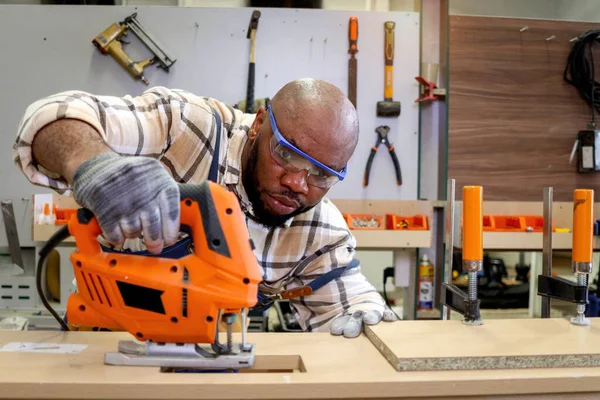 Middle-aged African carpenter wears safety glasses goggles and apron, using carpentry tools with wood plank to make wooden furniture in woodworking workshop, worker craftsman works at workshop.