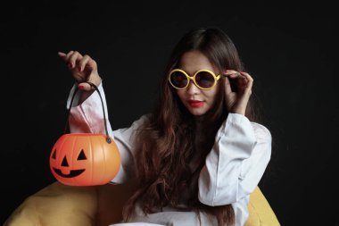 Beyaz hayalet kostümlü güzel çekici kadın turuncu balkabağı tutuyor, karanlık odada sarı koltukta oturuyor, makyajlı, korkunç yüzlü genç bayan Cadılar Bayramı partisini kutlamaya gidiyor..