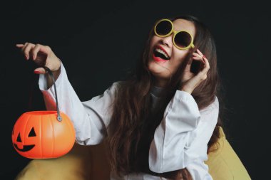 Beyaz hayalet kostümlü güzel çekici kadın turuncu balkabağı tutuyor, karanlık odada sarı koltukta oturuyor, makyajlı, korkunç yüzlü genç bayan Cadılar Bayramı partisini kutlamaya gidiyor..