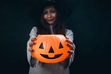 Cadılar Bayramı turuncu balkabağı elinde güzel siyah uzun saçlı ürkütücü beyaz hayalet kostümlü kadın karanlık odada duruyor makyajlı korkunç yüzlü kadın Cadılar Bayramı partisine gidiyor..