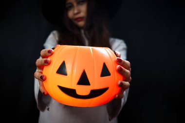 Cadılar Bayramı turuncu balkabağı elinde güzel siyah uzun saçlı ürkütücü beyaz hayalet kostümlü kadın karanlık odada duruyor makyajlı korkunç yüzlü kadın Cadılar Bayramı partisine gidiyor..