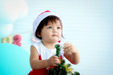 Noel Baba 'nın kırmızı kostümlü tatlı kız çocuğu beyaz oturma odasında Noel ağacı süslemeye çalışıyor, güzel küçük çocuk kış tatilini kutluyor, kutluyor, mutlu Noeller ve mutlu yıllar..