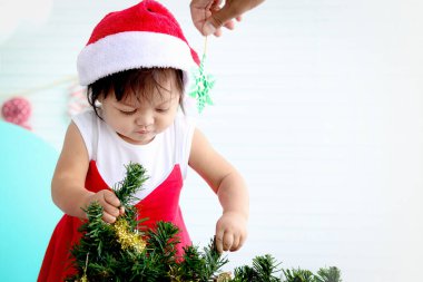 Noel Baba 'nın kırmızı kostümlü tatlı kız çocuğu beyaz oturma odasında Noel ağacı süslemeye çalışıyor, güzel küçük çocuk kış tatilini kutluyor, kutluyor, mutlu Noeller ve mutlu yıllar..