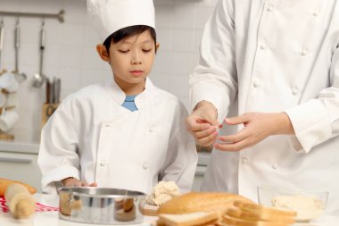 Mutlu Asyalı oğul ve aşçı üniformalı baba mutfakta birlikte yemek pişiriyorlar, baba ve çocuk ekmek hamuru hazırlıyorlar, şirin bir aile fırın yemeği yapıyor. Çocuk öğrenci ekmek pişirmeyi öğreniyor.