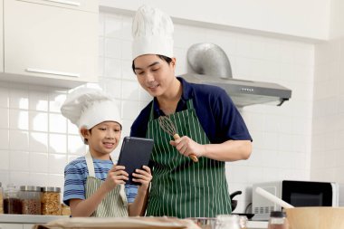 Mutlu gülümseyen Asyalı çocuk önlük takıyor ve şef şapkası takıyor, internette yemek tarifleri aramak için dijital tablet tutuyor, oğul ve baba mutfakta birlikte yemek pişiriyor, baba ve çocuk sevimli bir aile şefinde.