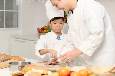 Mutlu Asyalı oğul ve aşçı üniformalı baba mutfakta birlikte yemek pişiriyorlar, baba ve çocuk ekmek hamuru hazırlıyorlar, şirin bir aile fırın yemeği yapıyor. Çocuk öğrenci ekmek pişirmeyi öğreniyor.